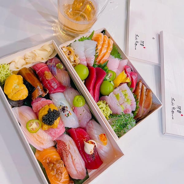Two Sushi Hatsune boxes filled with various colorful sushi and sashimi, with a drink beside them.